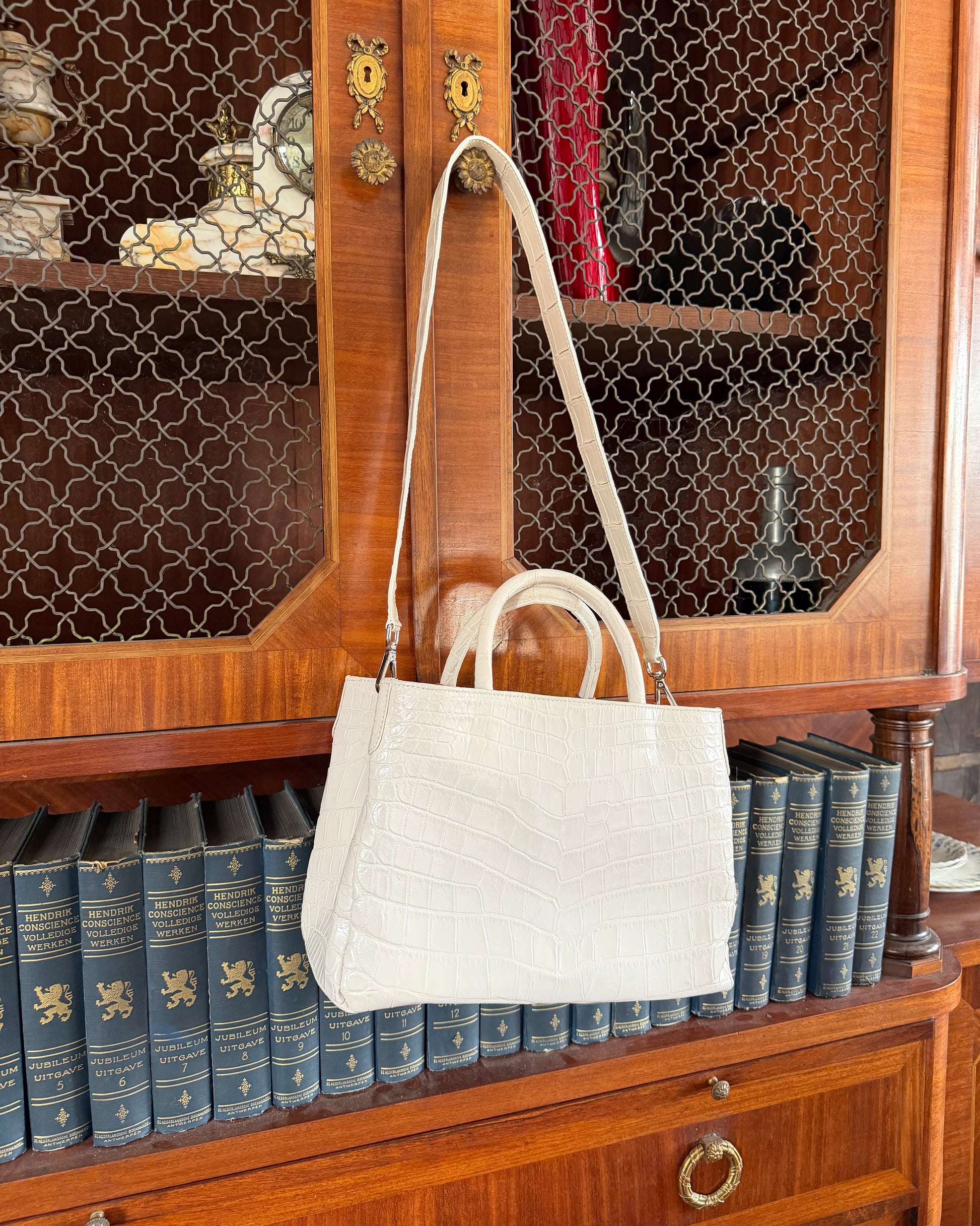 An off-white crocodile-embossed leather handbag with a single shoulder strap and top handles, hanging on a wooden cabinet door in front of a bookshelf.