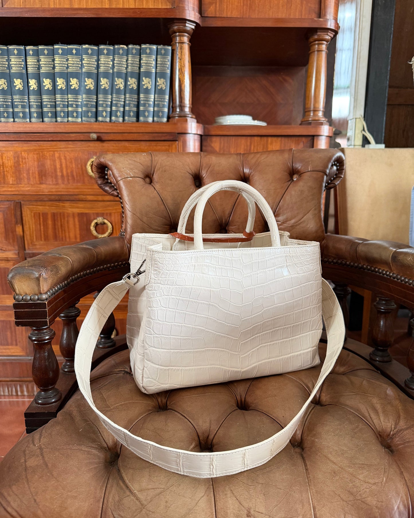 A structured beige crocodile-embossed leather handbag with double top handles and a matching shoulder strap, displayed on a brown leather tufted armchair in front of a wooden bookshelf.