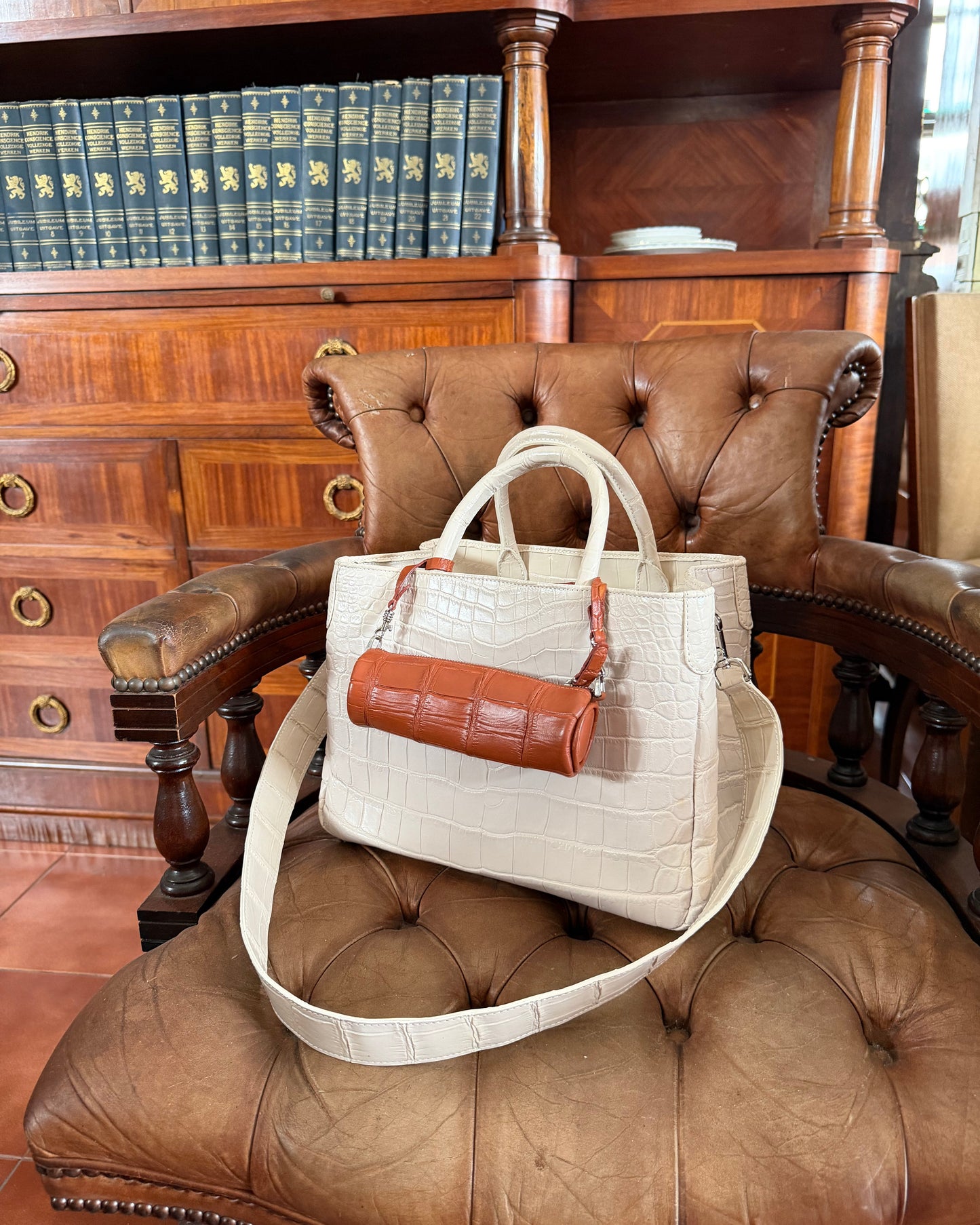 A structured handbag in off-white crocodile-embossed leather with a brown leather cylindrical pouch and matching strap, displayed on a brown tufted leather armchair in an indoor setting with wooden bookshelves.