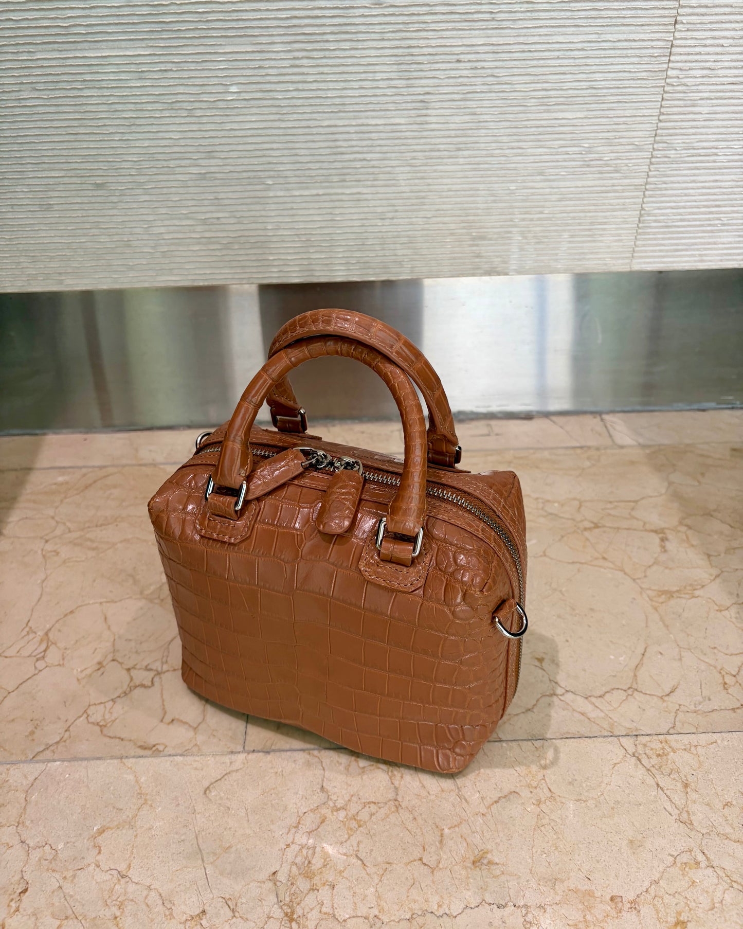 A brown crocodile-embossed leather handbag with double top handles and a zippered top, placed on a beige marble floor in front of a textured gray wall.