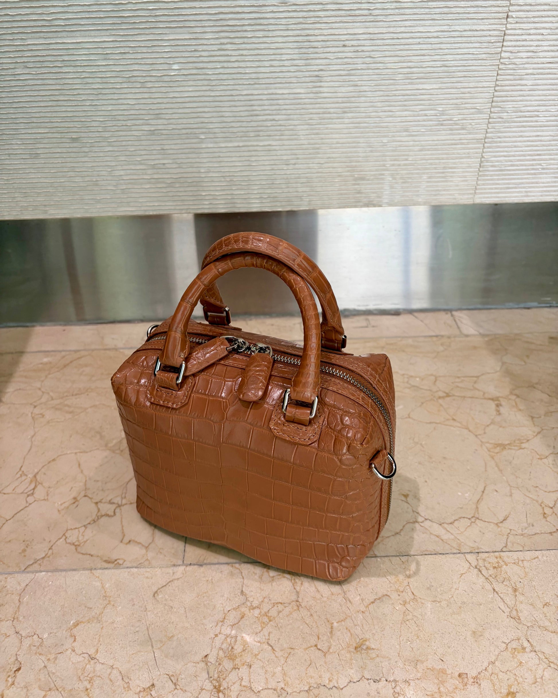 A brown crocodile-embossed leather handbag with double top handles and a zippered top, placed on a beige marble floor in front of a textured gray wall.