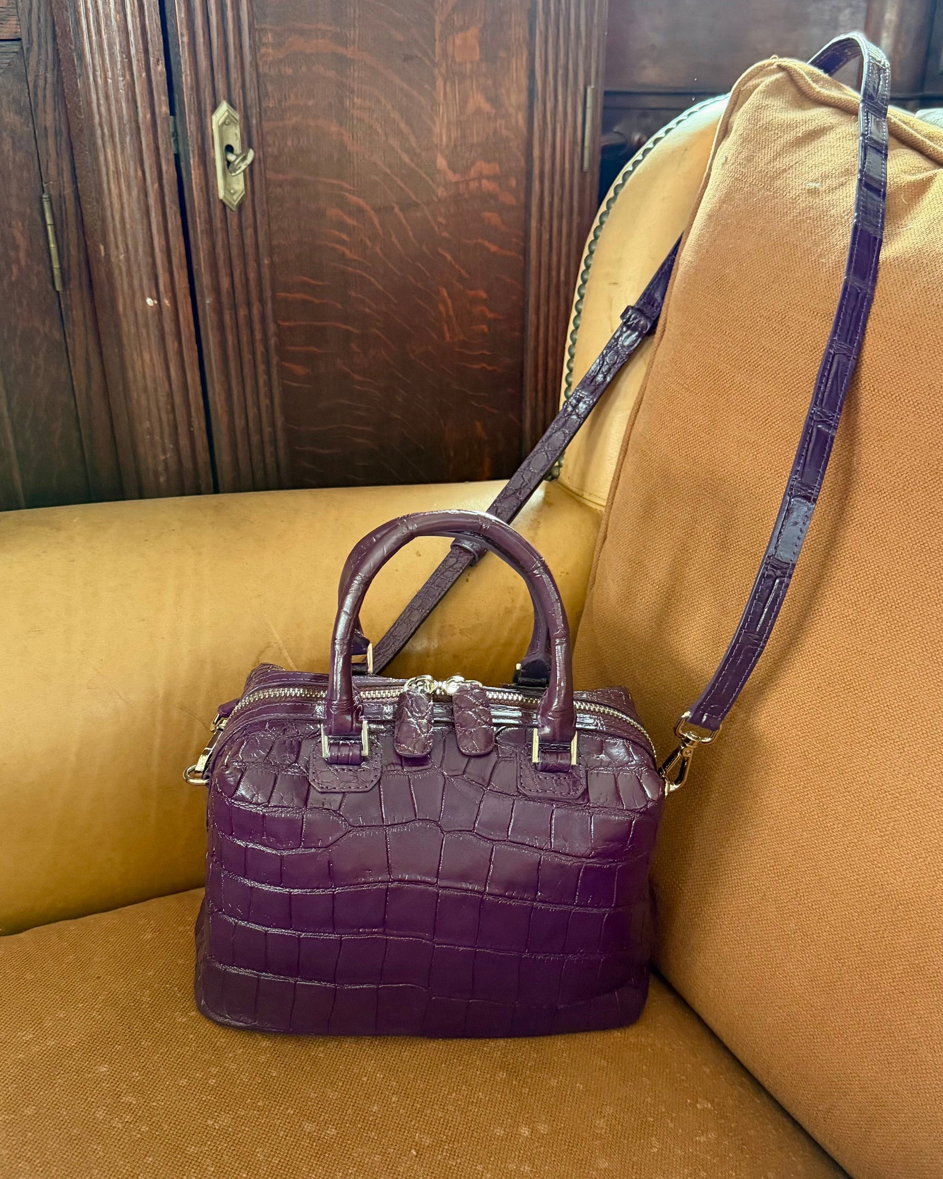 A purple crocodile-embossed leather handbag with double top handles and a matching shoulder strap, resting on a tan leather couch in front of a dark wood cabinet.