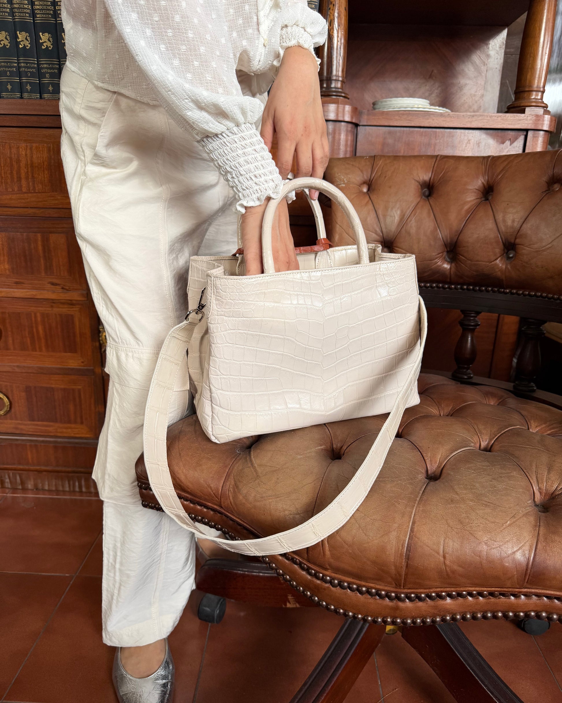 A person in a white patterned blouse and white trousers holds a structured cream crocodile-embossed handbag with double top handles and a long shoulder strap, standing beside a brown leather tufted chair in an indoor room.