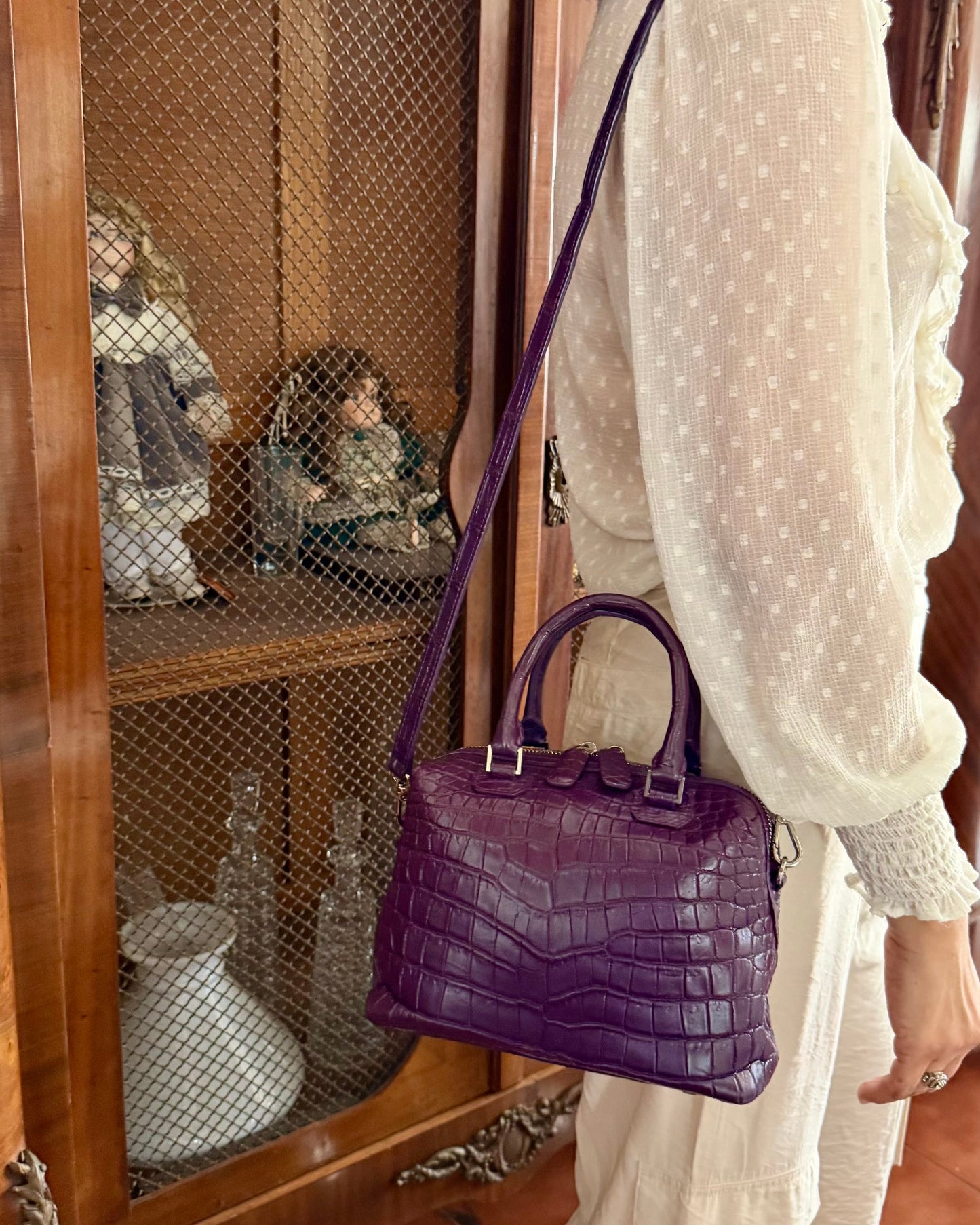 A woman wearing a white polka-dot blouse and white skirt carries a purple crocodile-embossed leather handbag with a shoulder strap, standing in front of a wooden display cabinet.