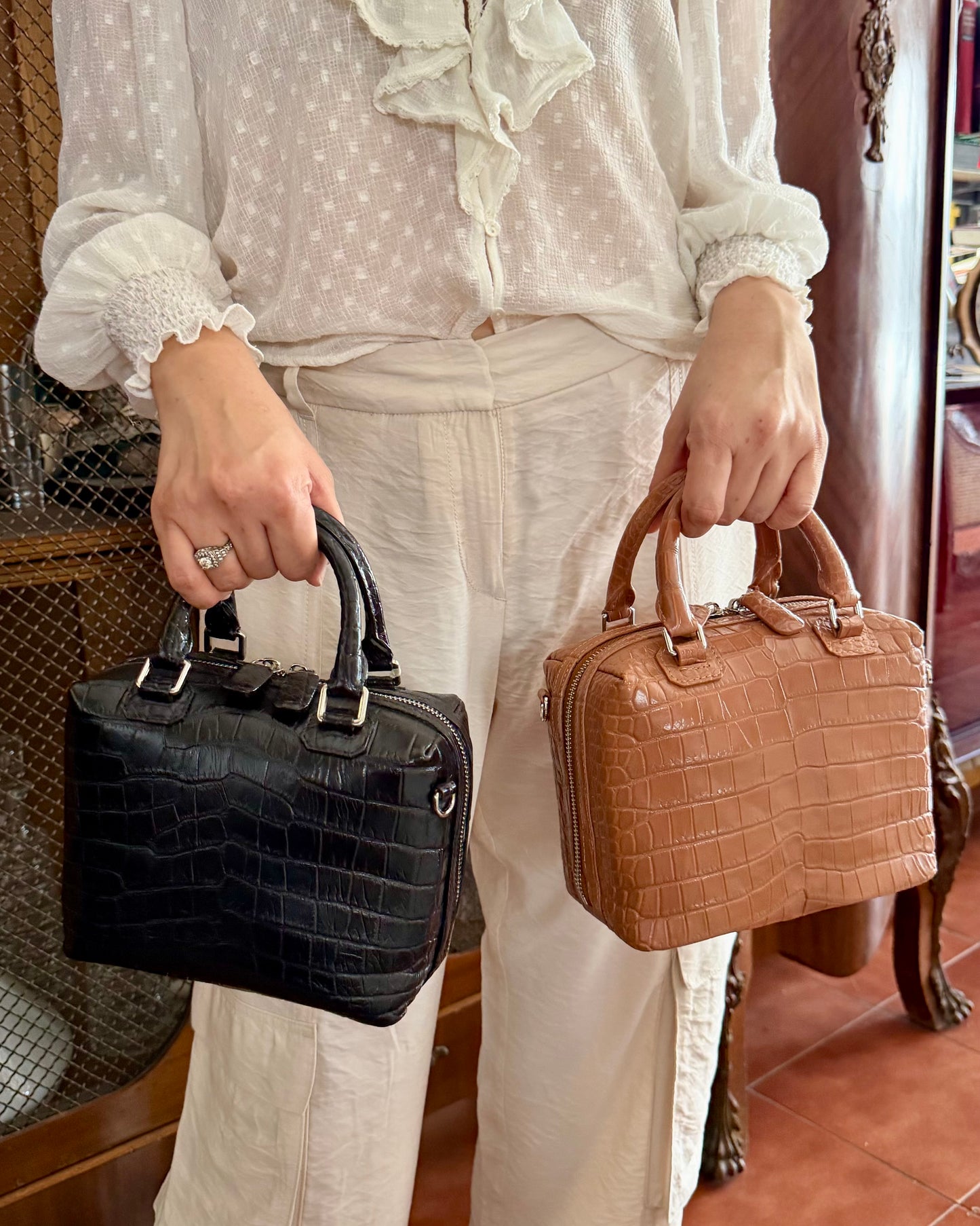 A person wearing a white blouse and cream pants holds two structured top-handle handbags, one black crocodile-embossed and one tan crocodile-embossed, in an indoor setting.