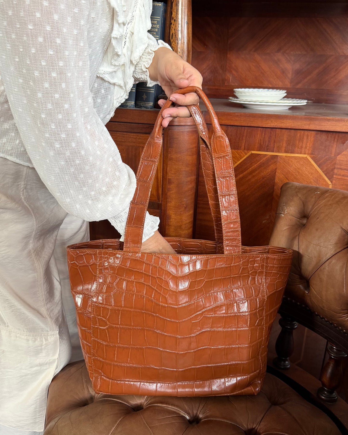 A person in a white polka-dot blouse holds a large brown crocodile-embossed tote bag in a warm, rustic indoor setting with a wooden cabinet and tufted leather chair.