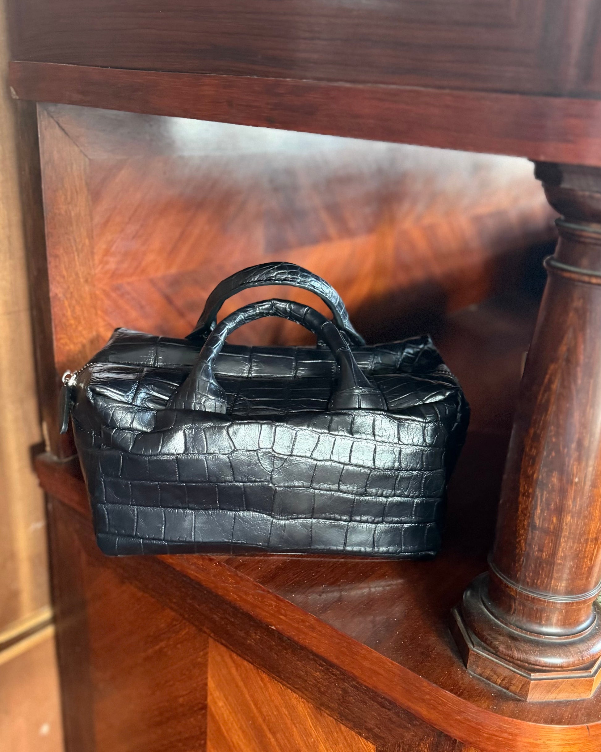 A small black crocodile-embossed leather handbag with double top handles, resting on a wooden shelf next to a wooden column.