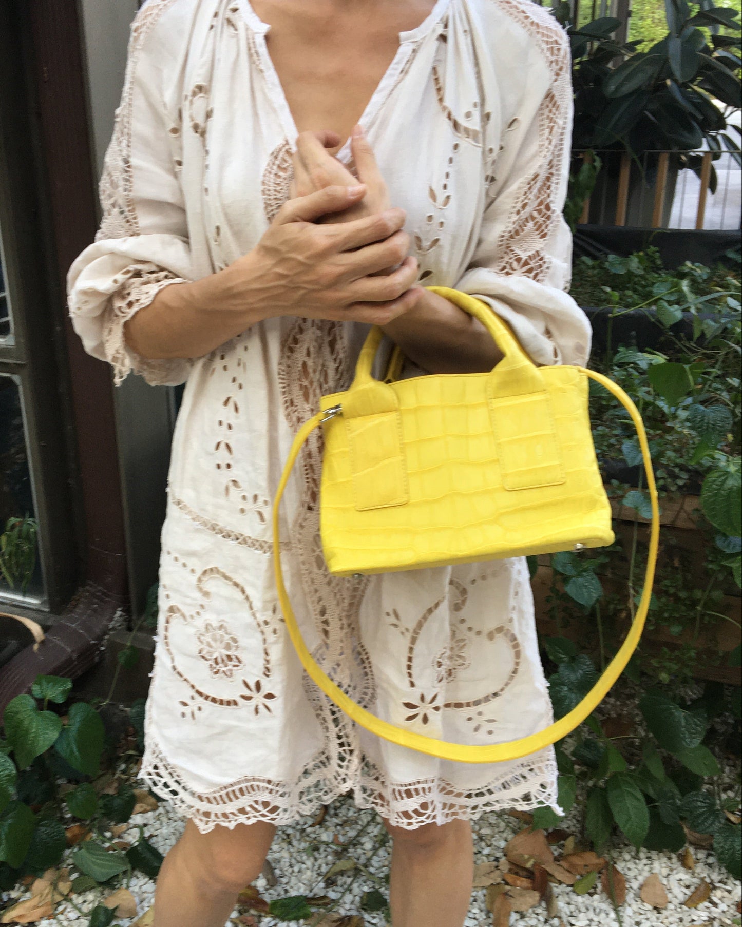 Person holding a yellow handbag with a white lace dress and greenery in the background