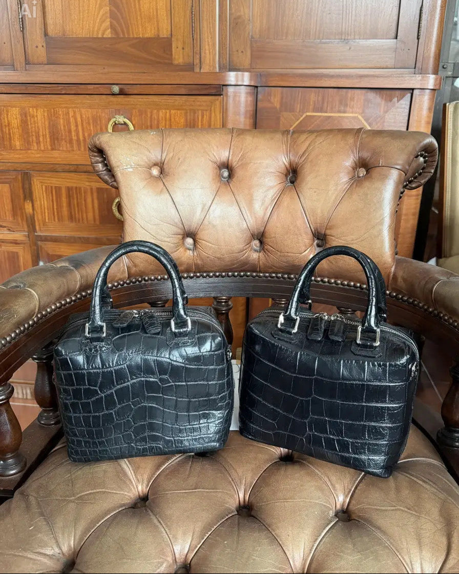 Two structured black crocodile-embossed handbags with double top handles, displayed side-by-side on a brown leather tufted armchair in front of a wooden cabinet.