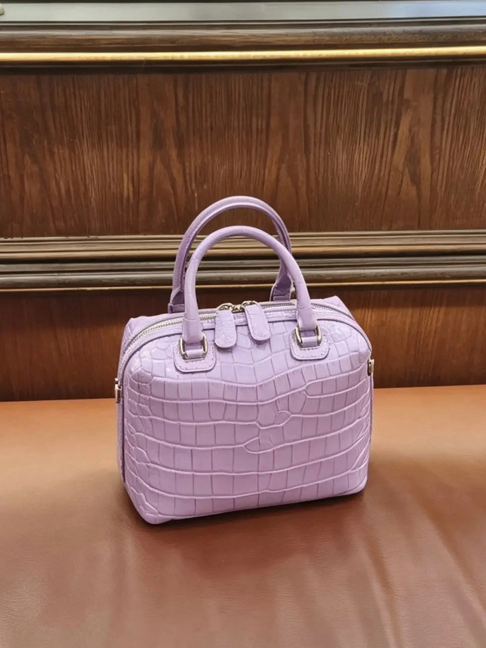 A light purple crocodile-embossed leather handbag with double top handles and gold-tone hardware, resting on a brown leather surface against a dark wood-paneled background.