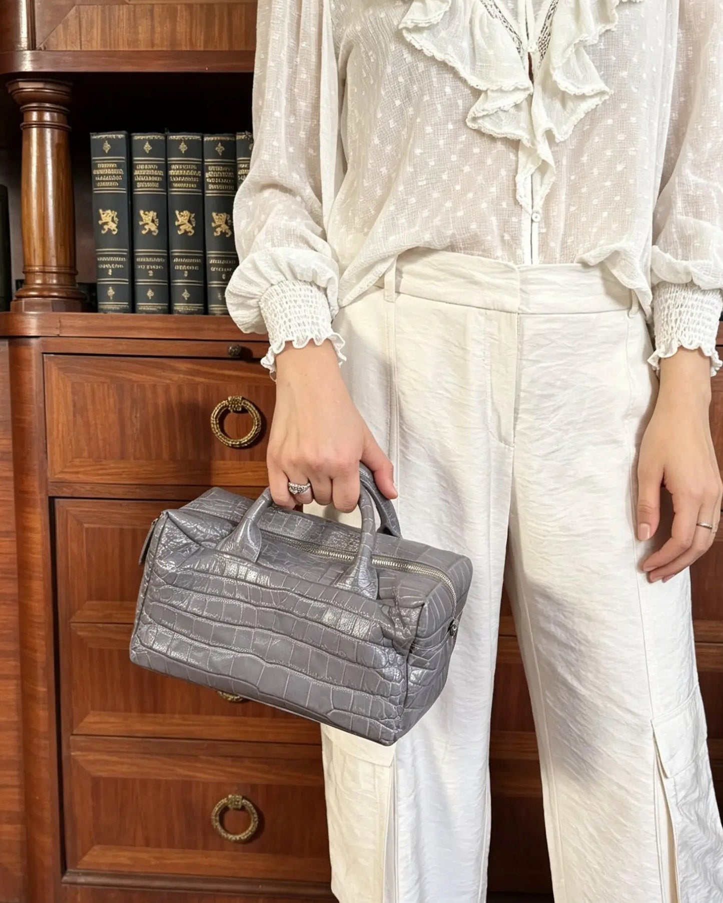 A person wearing a white ruffled blouse and white wide-leg trousers stands in front of a wooden bookcase, holding a gray crocodile-embossed handbag.