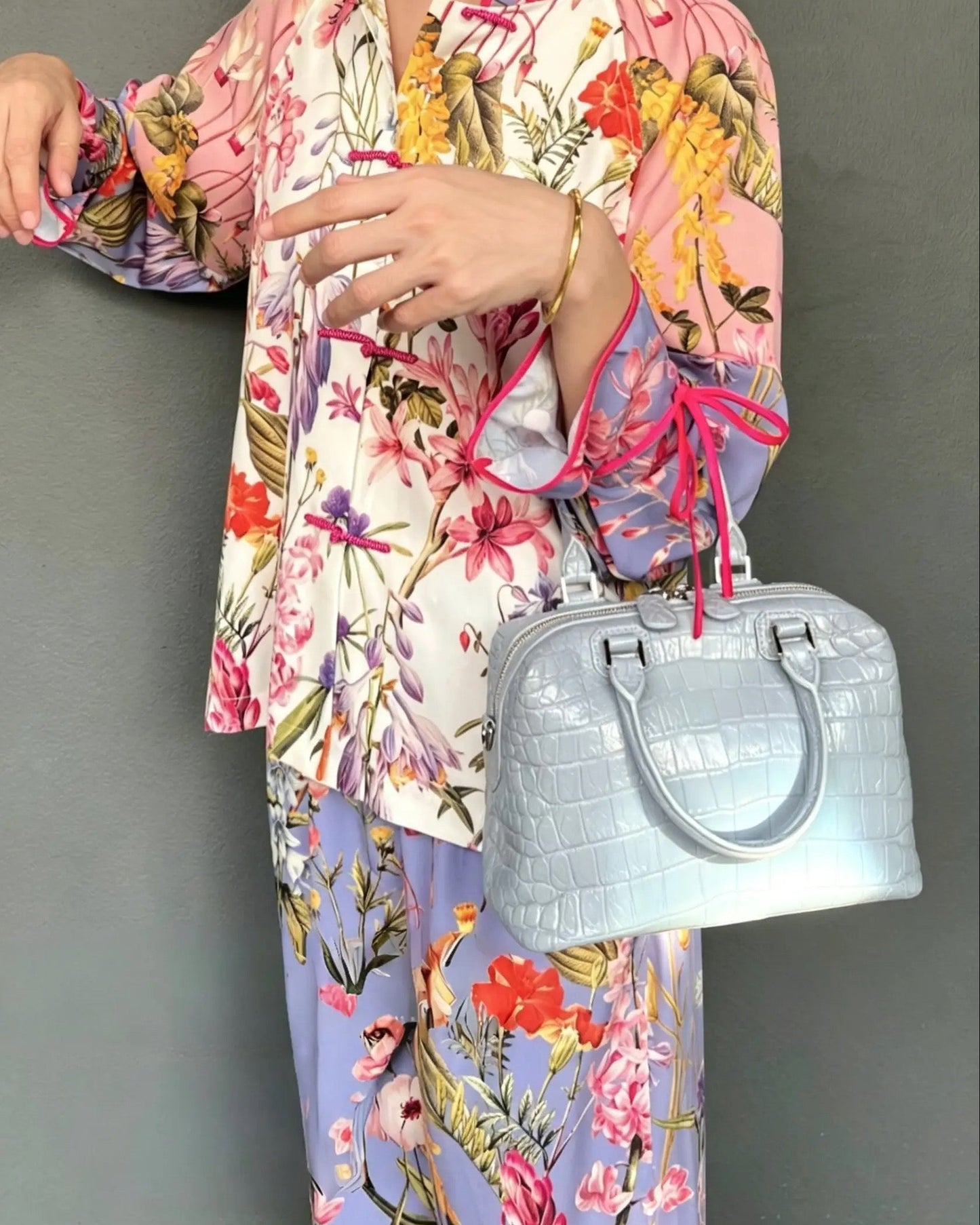 A person wearing a matching floral pajama set with pink and lavender tones, holding a light blue crocodile-embossed handbag against a plain gray wall.