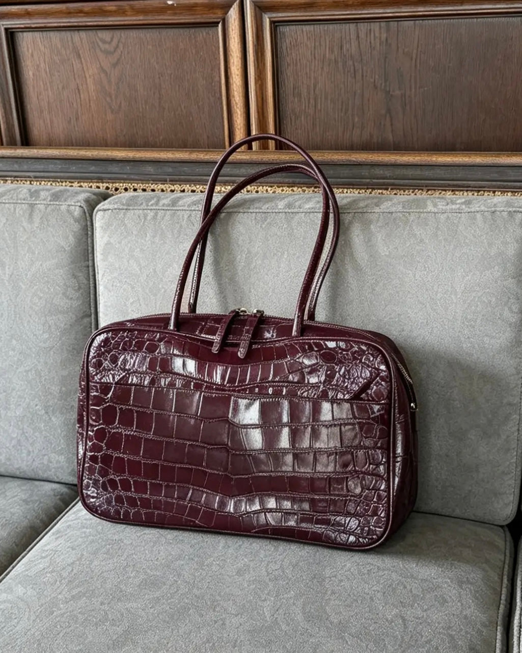 A dark burgundy crocodile-embossed leather handbag with double top handles, resting on a gray upholstered sofa in front of a wooden paneled wall.