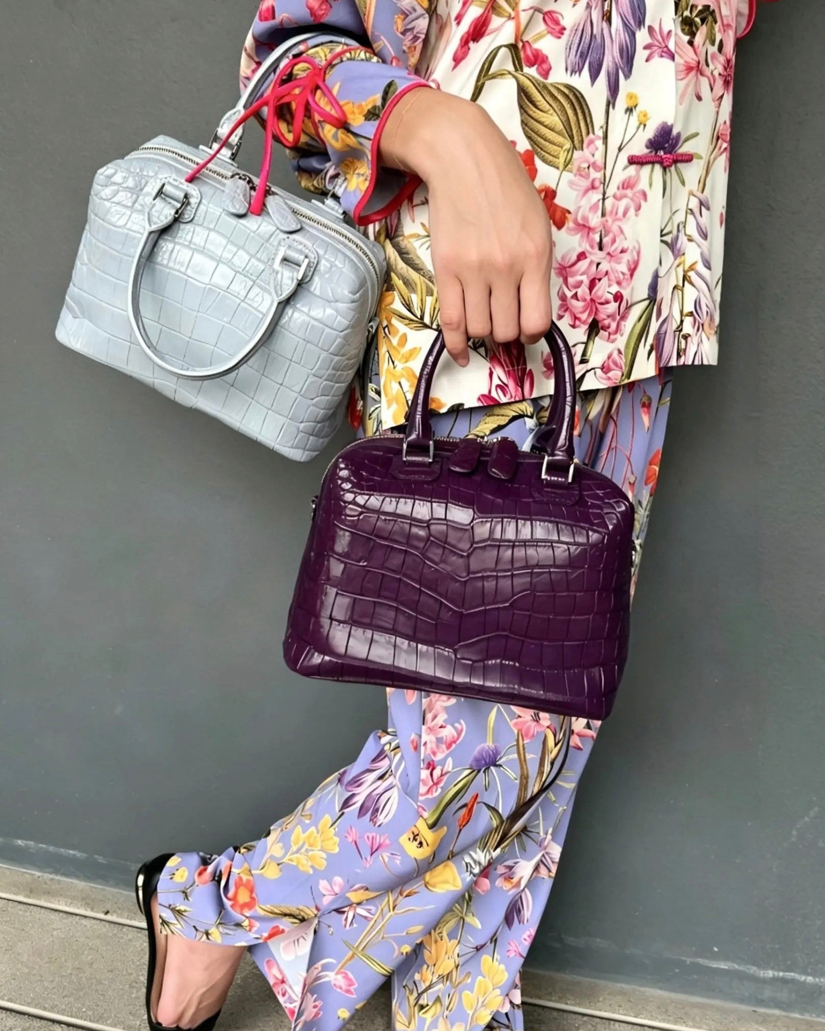A person wearing a colorful floral print outfit holds two crocodile-embossed handbags against a gray wall. One bag is light silver-gray, and the other is deep purple.