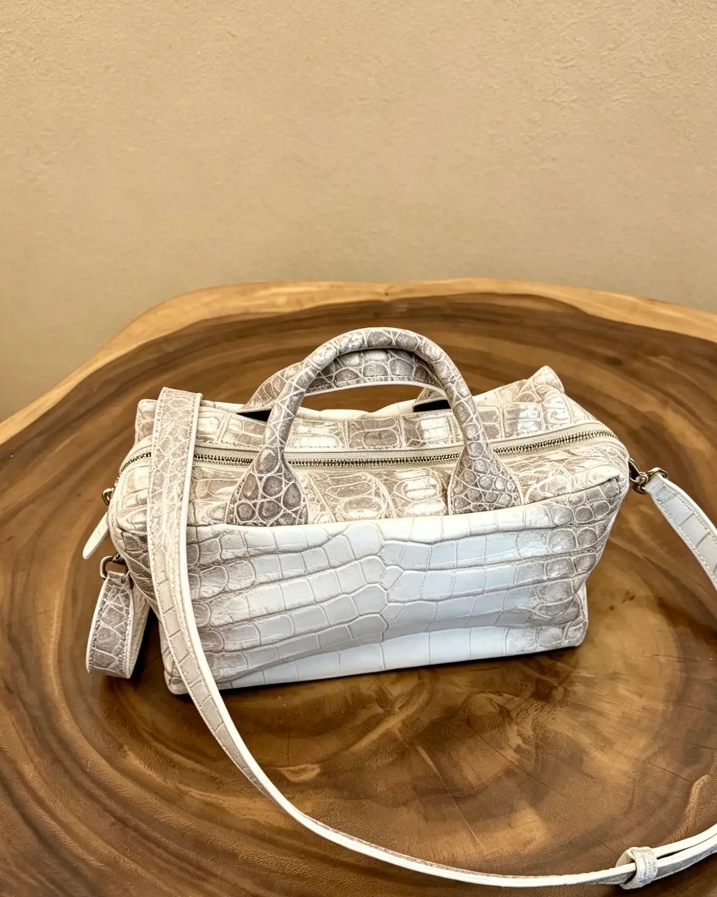A light beige and white crocodile-embossed leather handbag with dual top handles and a matching shoulder strap, displayed on a round wooden slice against a beige wall.