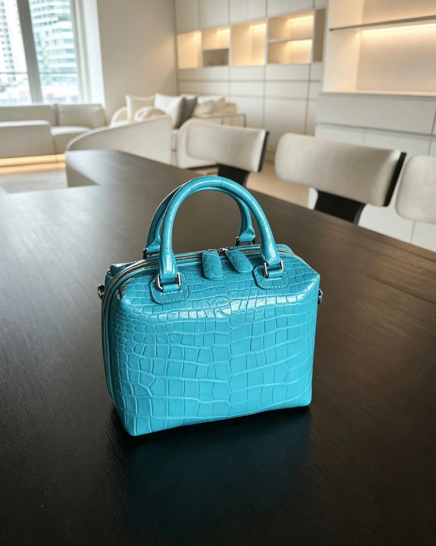 A small turquoise crocodile-embossed handbag with double top handles, placed on a dark wooden table in a modern, brightly lit living room.