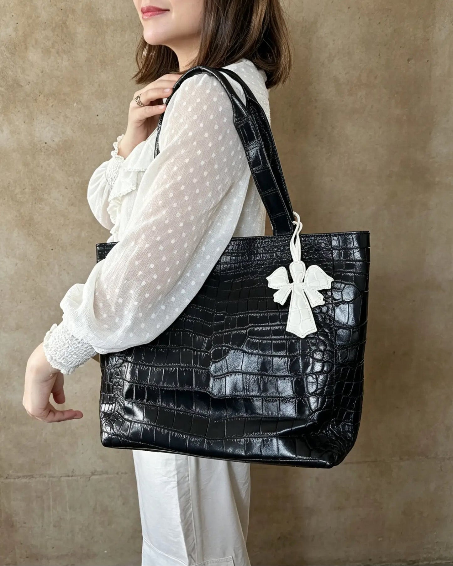 A woman wearing round sunglasses and a white polka-dot blouse carries a large black crocodile-embossed tote bag with a white bow detail against a textured beige wall.