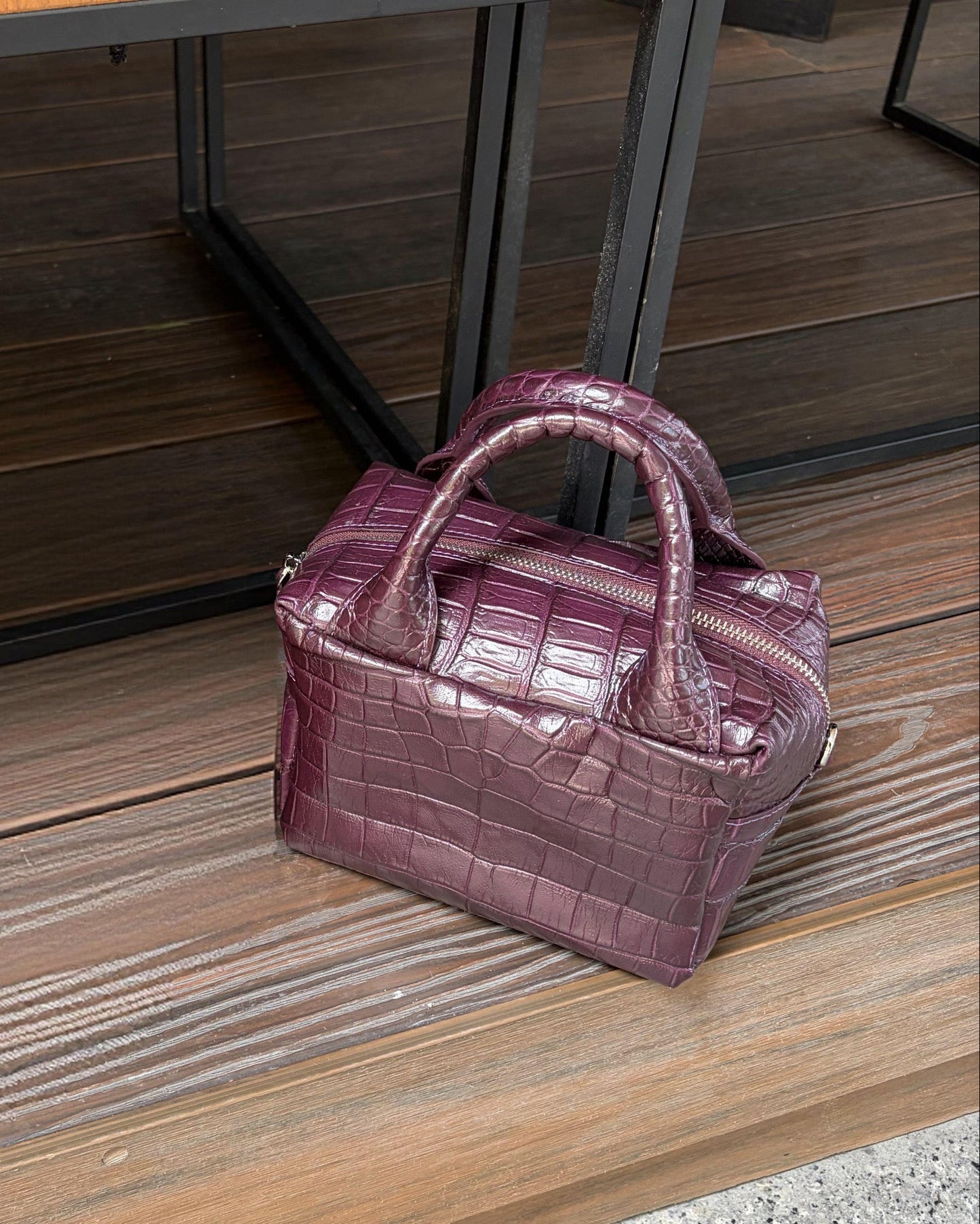 A small structured handbag with a glossy crocodile-embossed texture in a muted plum-purple color, resting on a wooden deck next to a glass table.