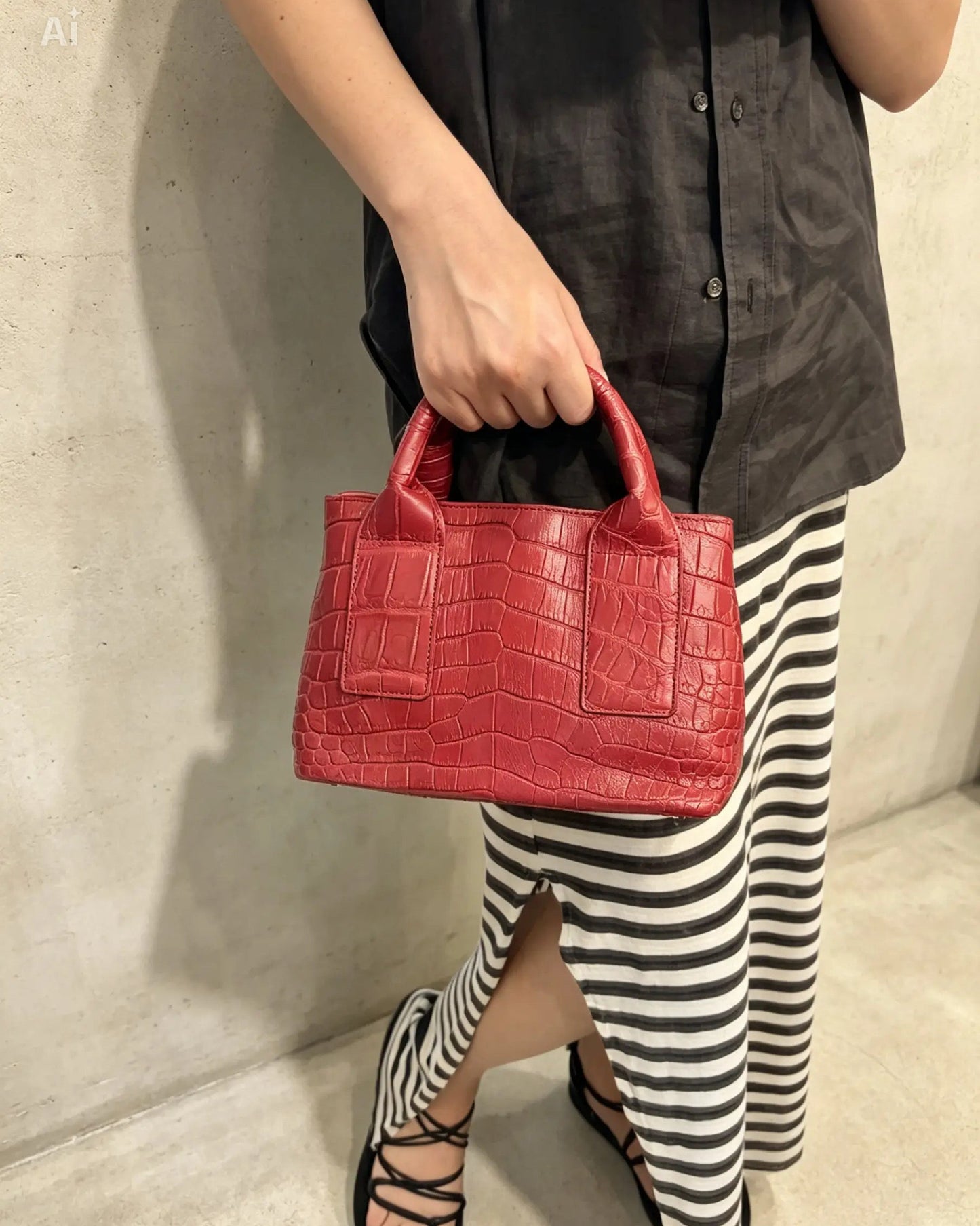 A person wearing a black button-up shirt and black-and-white striped skirt holds a bright red crocodile-embossed leather handbag with top handles, standing against a light concrete wall.
