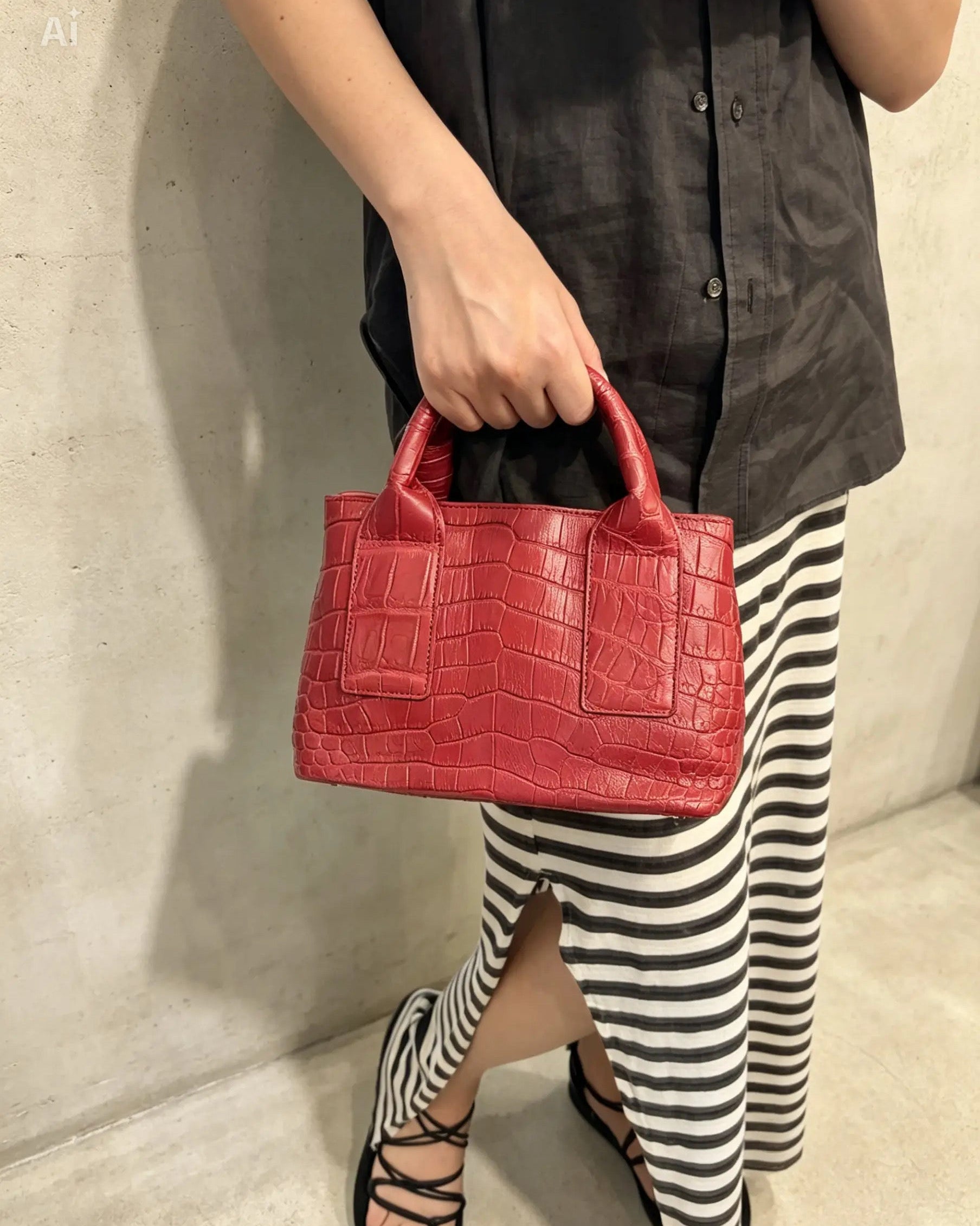 A person wearing a black button-up shirt and black-and-white striped skirt holds a bright red crocodile-embossed leather handbag with top handles, standing against a light concrete wall.