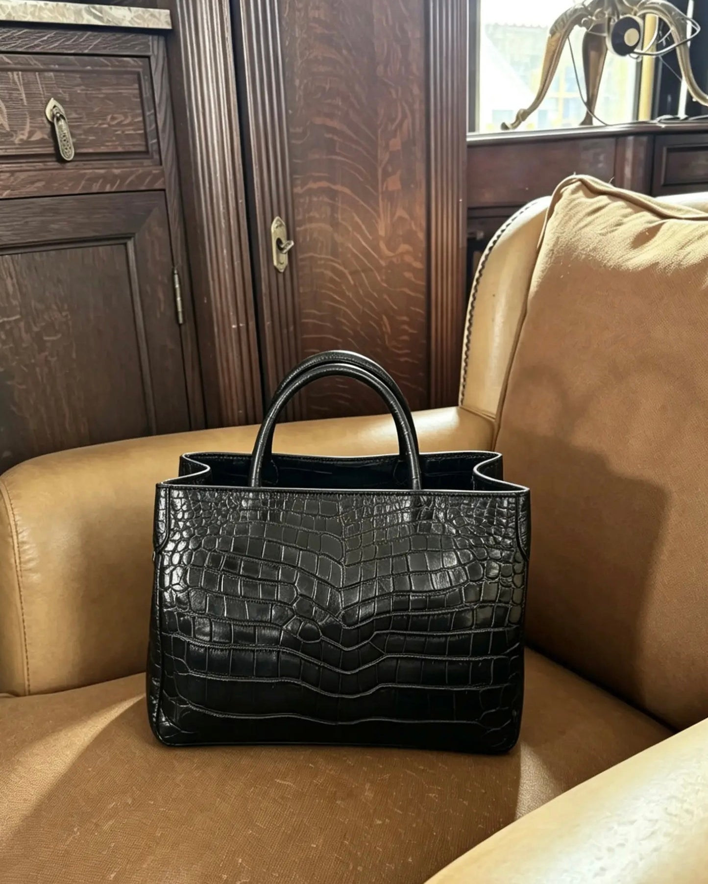 A black crocodile-embossed leather handbag with double top handles, resting on a tan leather armchair in front of a dark wood cabinet.