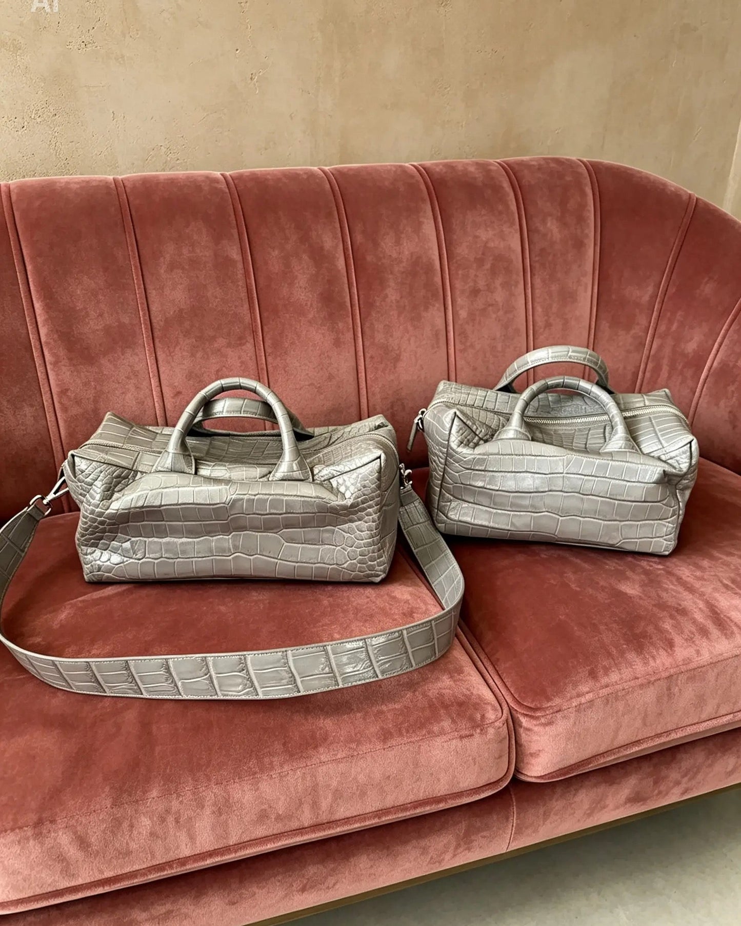 Two metallic silver crocodile-embossed handbags with top handles and detachable straps, displayed on a dusty rose velvet sofa against a beige wall.