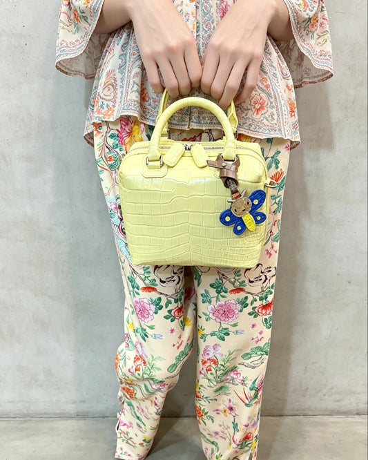 A person wearing a floral print top and matching pants holds a light yellow crocodile-embossed handbag with a blue flower charm against a gray concrete wall.