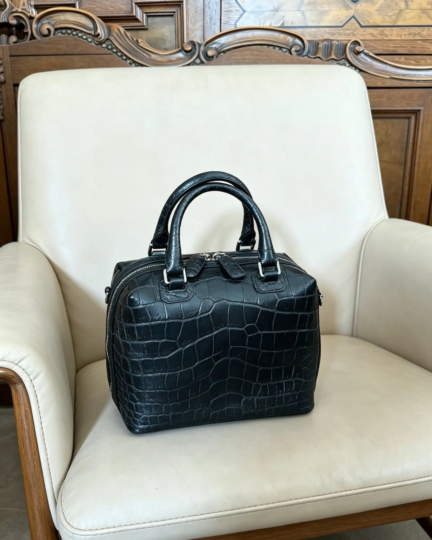 A black crocodile-embossed leather handbag with double top handles, resting on a cream-colored leather armchair in front of a wooden cabinet.
