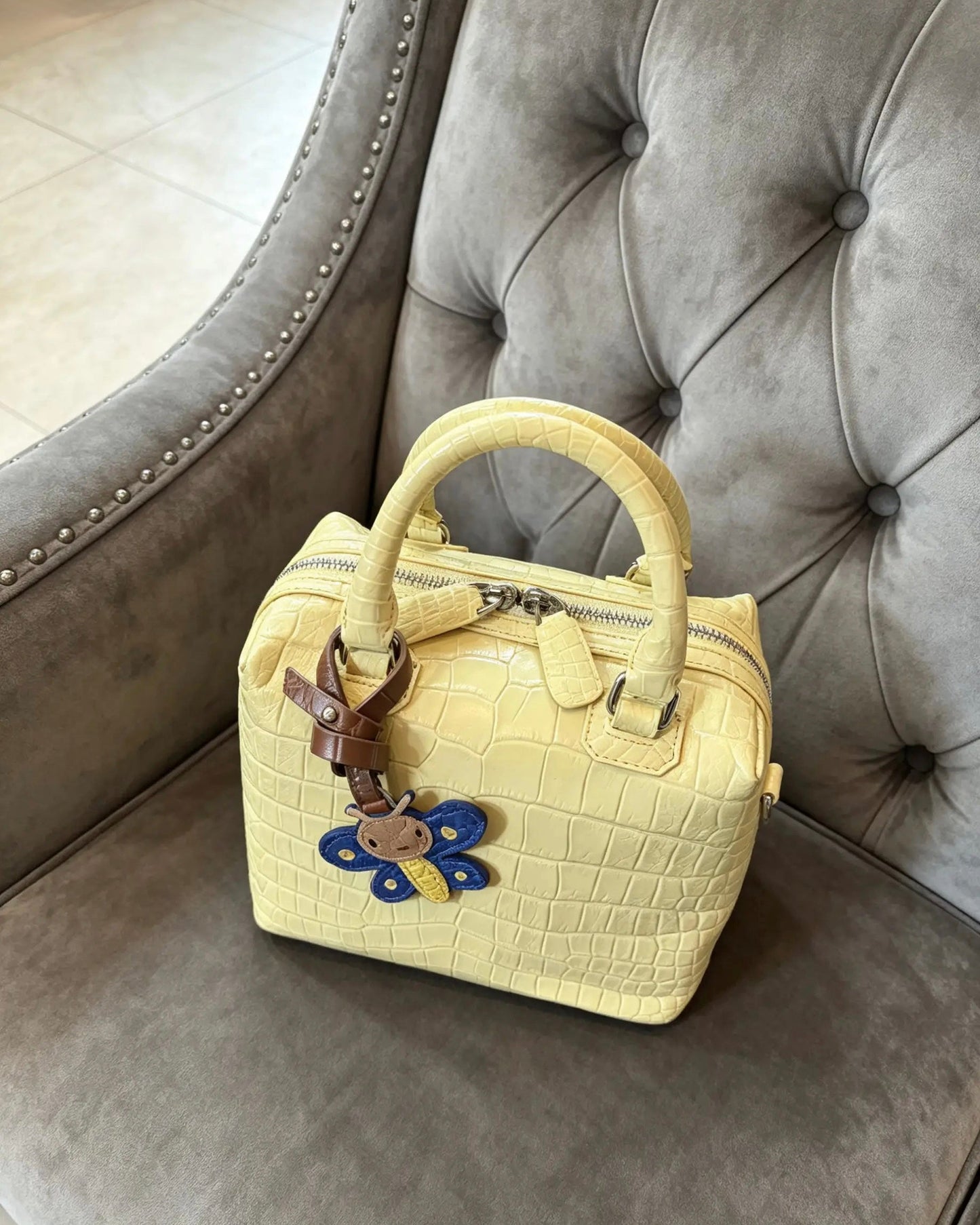 A pale yellow crocodile-embossed leather handbag with dual top handles and a decorative blue and yellow flower charm, resting on a gray tufted armchair.