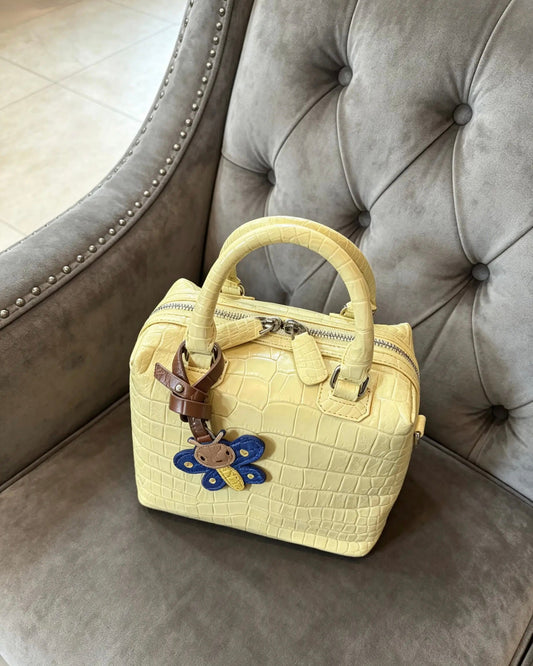A pale yellow crocodile-embossed leather handbag with dual top handles and a decorative blue and yellow flower charm, resting on a gray tufted armchair.
