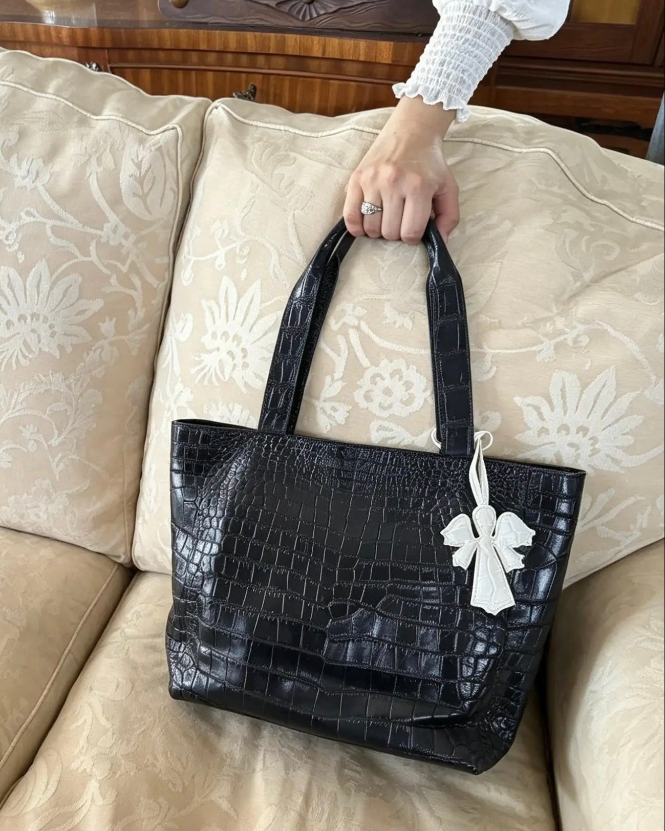 A black crocodile-embossed leather tote bag with double top handles, held by a person's hand on a beige patterned sofa. The bag features a white fabric bow tag hanging from one handle.