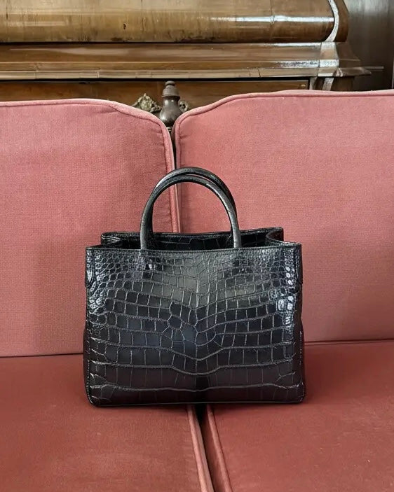 A black crocodile-embossed leather handbag with double top handles, resting on a dusty pink upholstered bench.