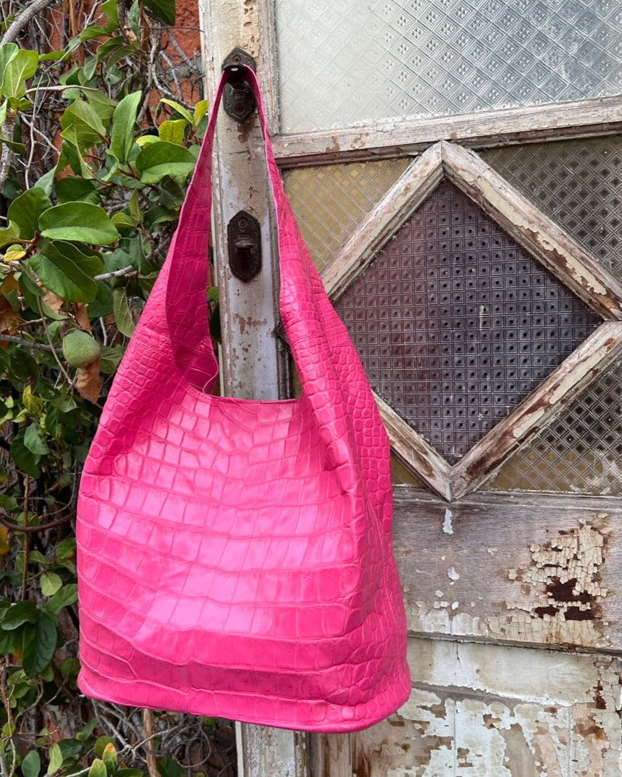 A fuchsia pink crocodile leather hobo bag with a magnet closure, hanging against an old wooden background door.
