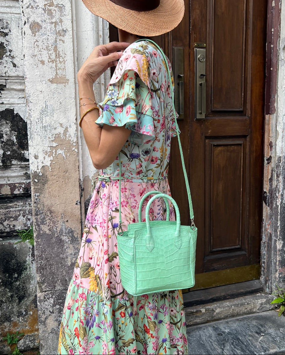Woman in a floral dress and straw hat holding a green handbag, standing in front of a wooden door.