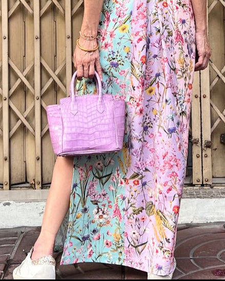 Woman in a floral dress and straw hat holding a pink bag, standing in front of a wooden lattice fence.