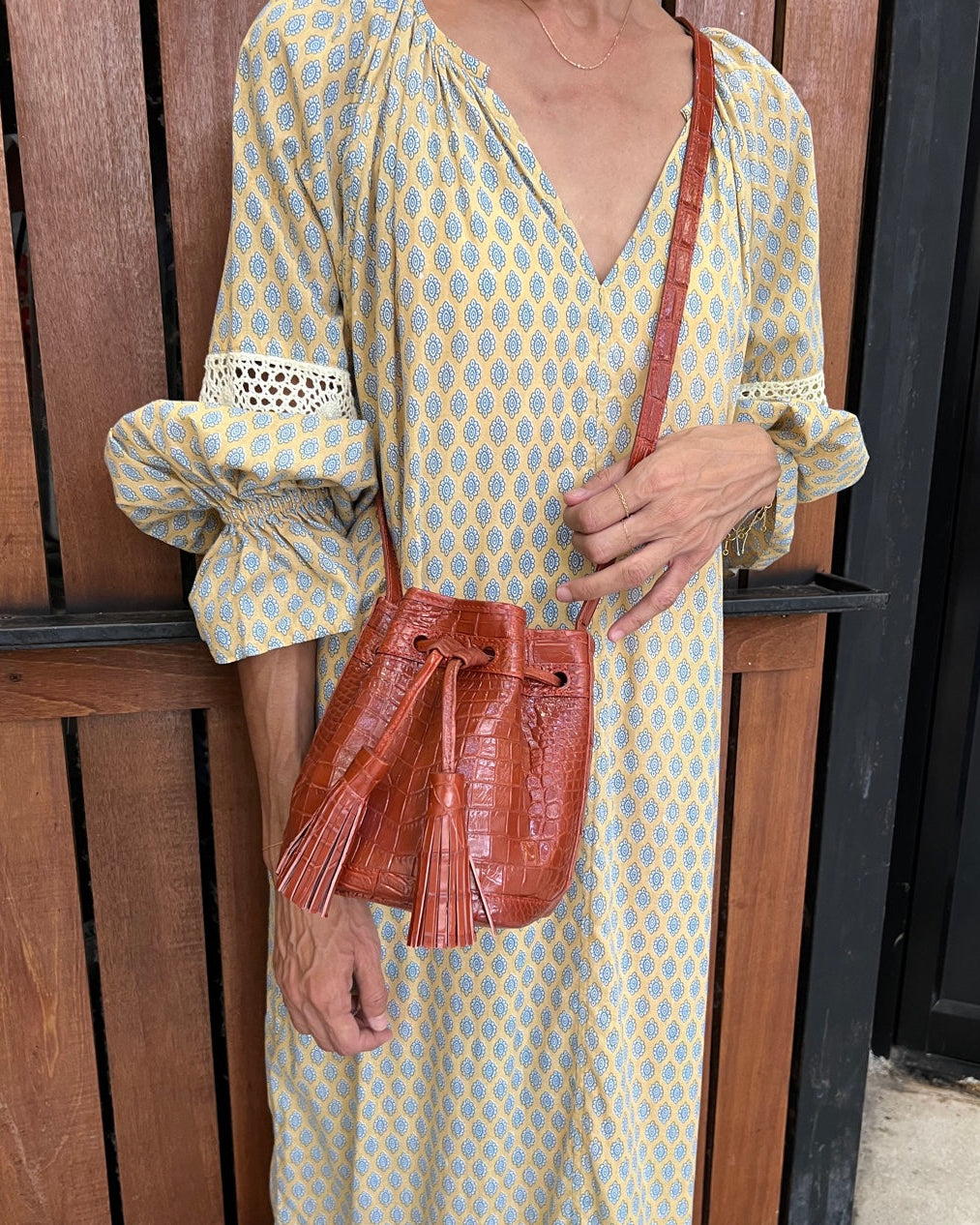 A woman wearing a light yellow and blue patterned maxi dress with lace trim, holding a brown crocodile-embossed crossbody bag with tassel details.