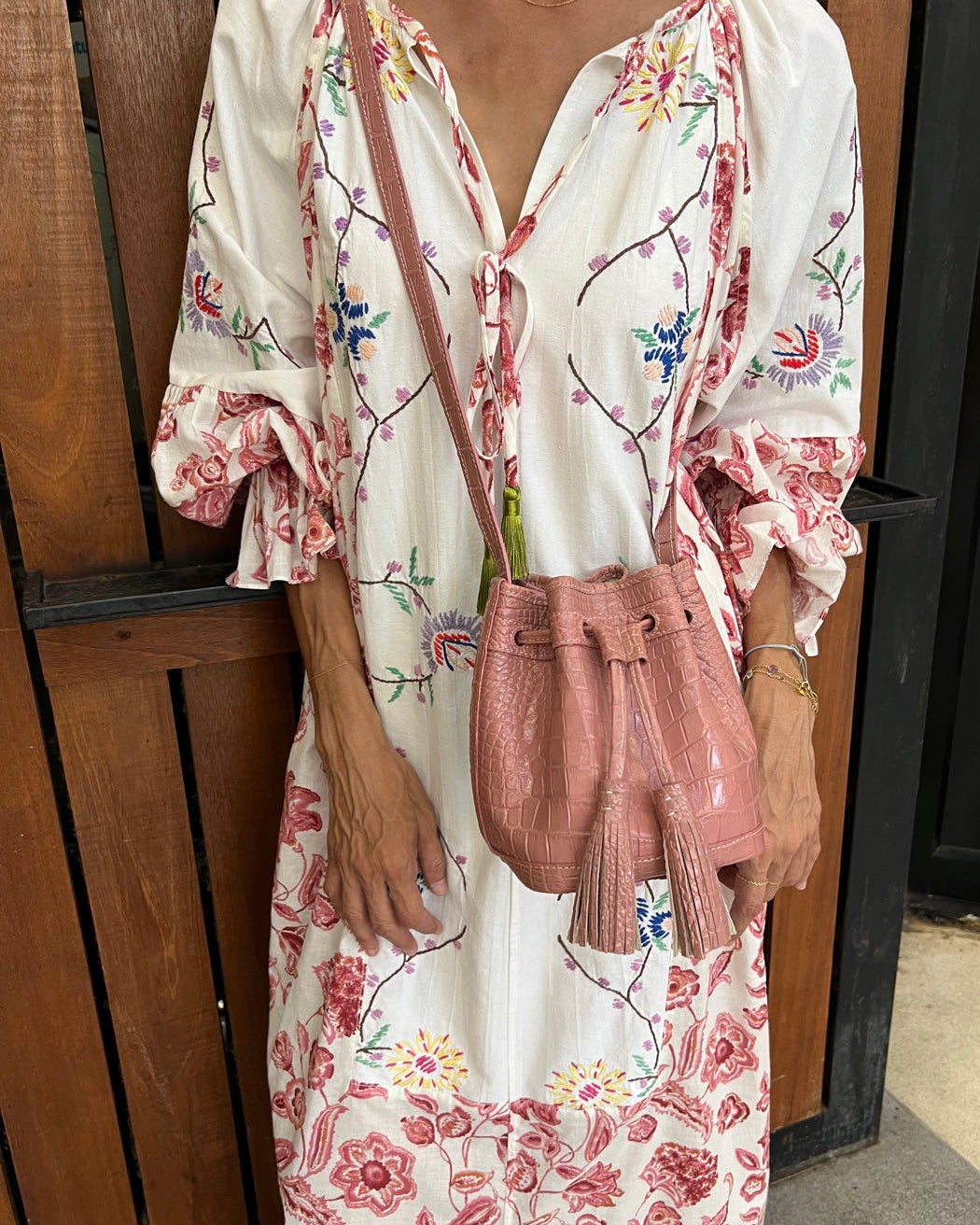 A woman wearing a white long-sleeve dress with a pink floral print and carrying a pink croc-embossed crossbody bag with tassels, standing in front of a wooden door.