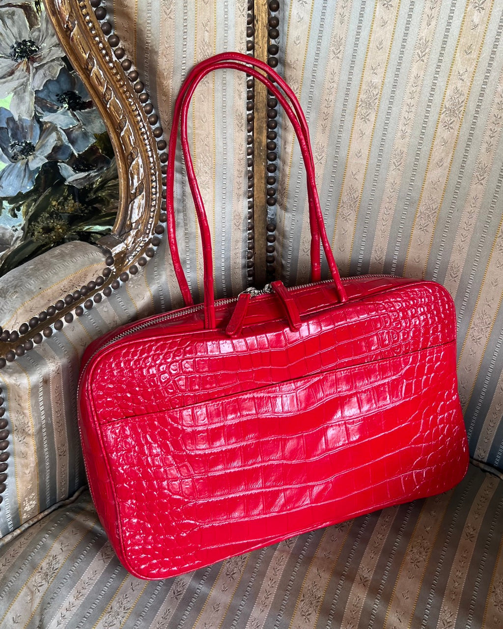 A bright red crocodile-embossed leather handbag with double top handles, resting on a vintage patterned armchair.